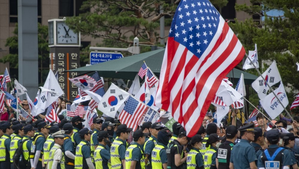A crowd of people are waving U.S. and South Korean flags, standing behind a line of policemen.